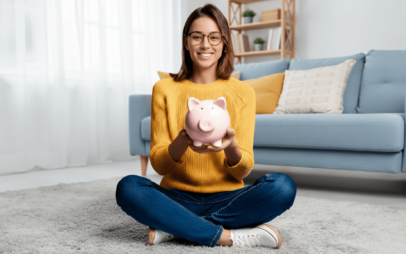 Una ragazza sorridente con gli occhiali e un maglione giallo, seduta a gambe incrociate sul pavimento di un salotto, mentre tiene in mano un salvadanaio rosa.