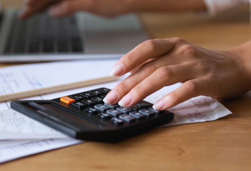 Mano di una persona che utilizza una calcolatrice su una scrivania con documenti e un laptop sullo sfondo, che simboleggia il calcolo del coefficiente di trasformazione per la conversione del montante in rendita.