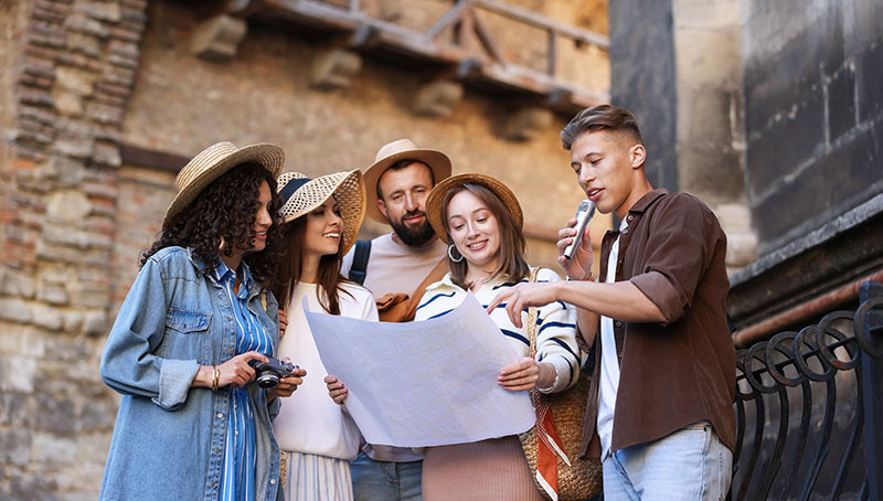 Una guida turistica, figura che rappresenta i lavoratori stagionali, mostra una mappa cartacea a un gruppo di turisti durante una visita guidata all'aperto vicino a un edificio storico.