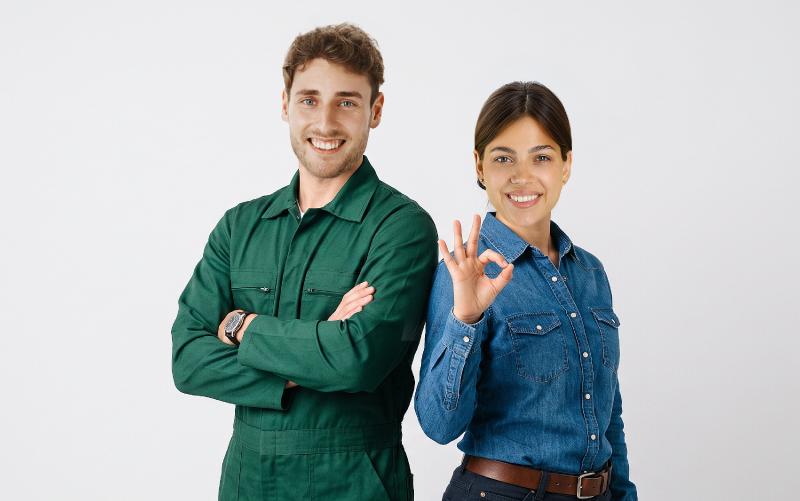 Un uomo in tuta da lavoro verde con le braccia conserte e una donna in camicia di jeans che fa il segno dell'OK con la mano, entrambi sorridenti su sfondo bianco." Opzione breve: alt="Due lavoratori sorridenti, un uomo in tuta verde e una donna che fa il gesto dell'OK.
