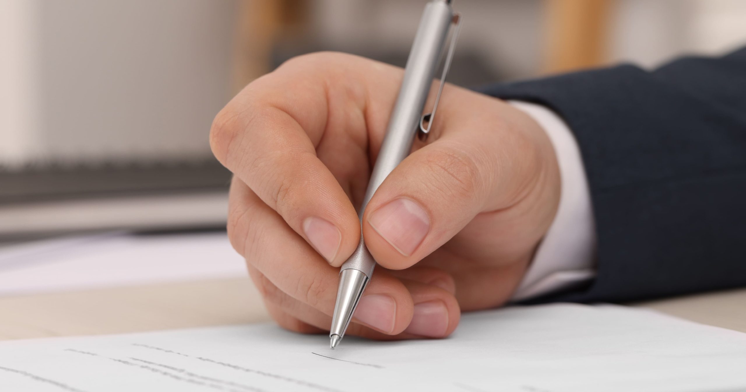 Primo piano della mano di una persona in giacca scura che firma un documento istituzionale con una penna a sfera argentata.
